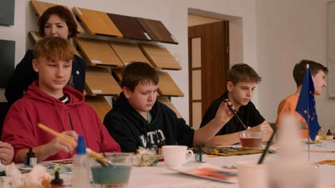 A group of boys sits at a table engaged in painting activities, with art supplies scattered around. A woman stands in the background, observing. Paintings and color samples are displayed on the wall behind them.