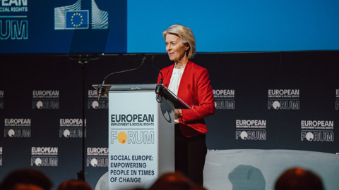 A woman in a red blazer stands at a podium labeled "European Forum" and "Social Europe: Empowering People in Times of Change," speaking at an event. The background features forum branding and an EU flag graphic.