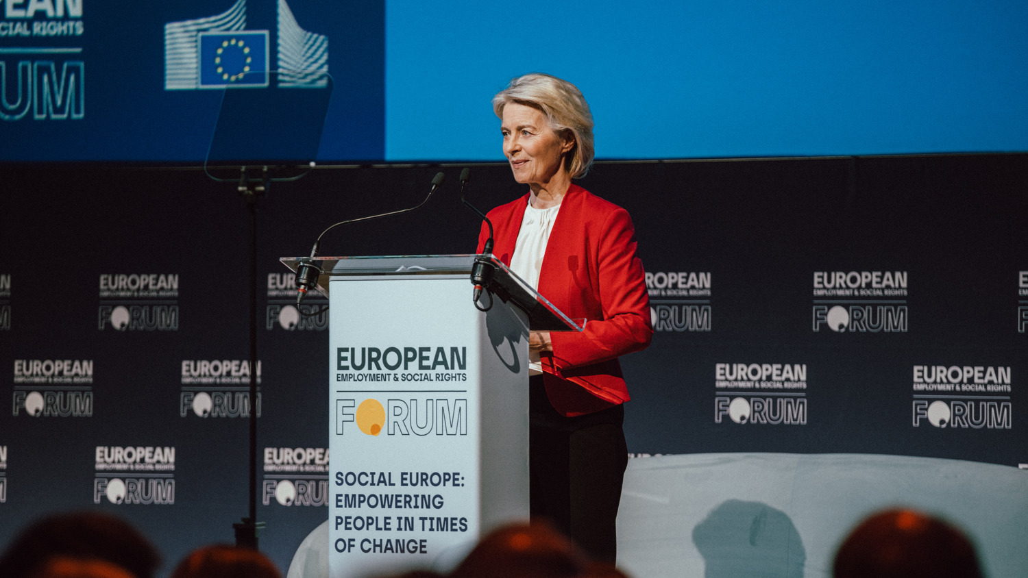 A woman in a red blazer stands at a podium labeled "European Forum" and "Social Europe: Empowering People in Times of Change," speaking at an event. The background features forum branding and an EU flag graphic.
