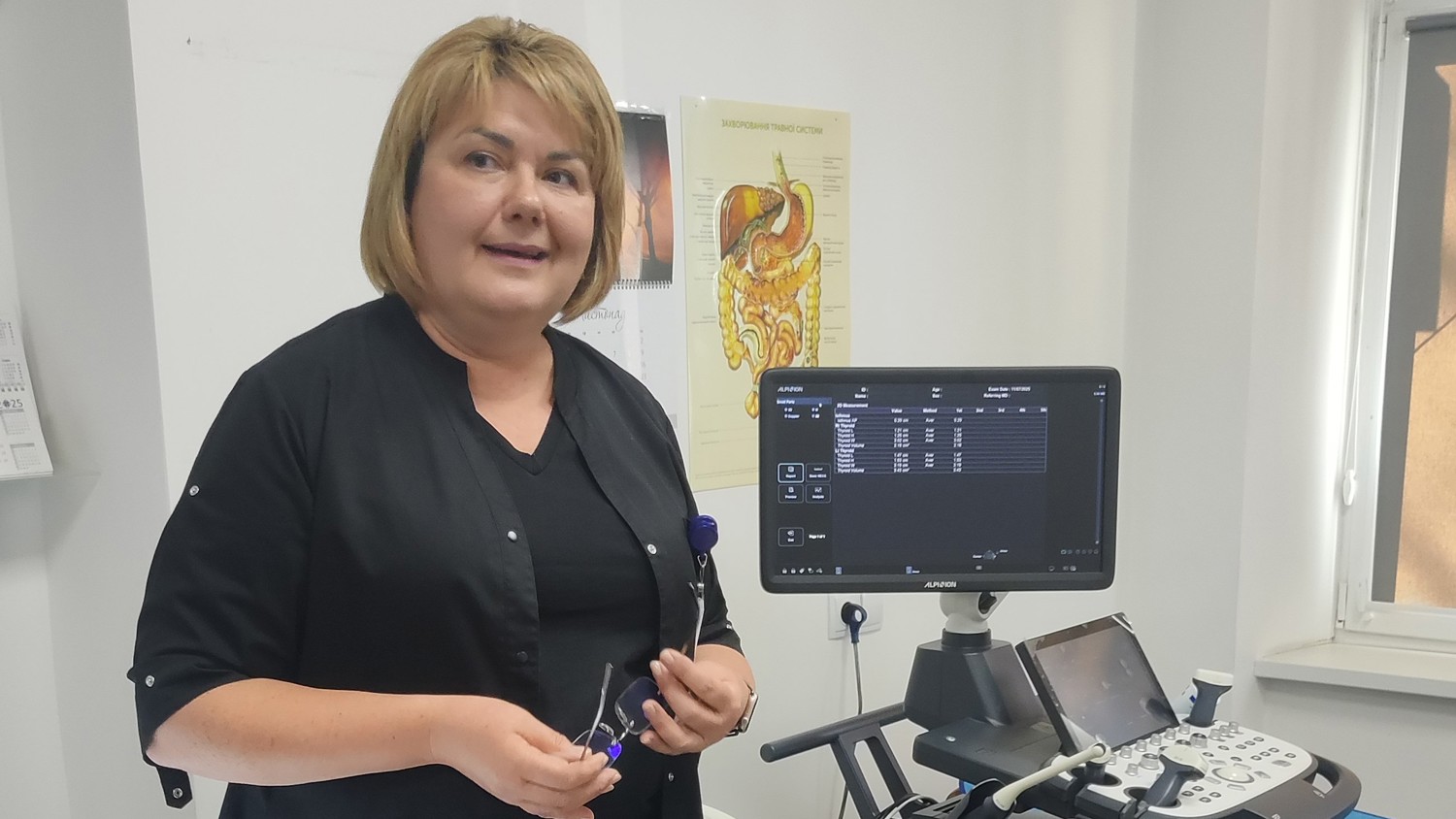 A woman in a black coat stands in a medical office beside an ultrasound machine, holding a medical tool. Medical diagrams and papers are visible on the wall and desk around her.