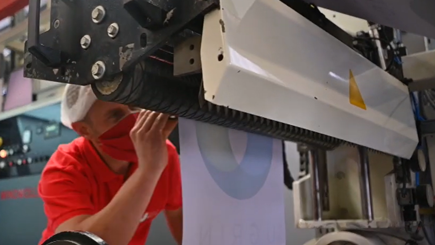 A person wearing a red shirt and red mask inspects a large printing machine. The machine is processing a roll of paper labeled "ONYTACK." The background shows parts of the industrial equipment.
