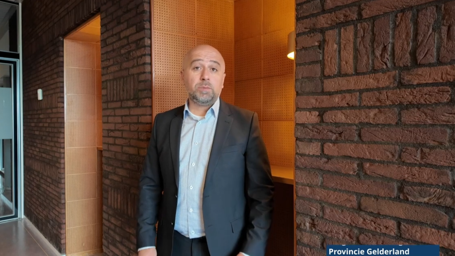A man in a suit stands indoors near a brick wall, with a wooden alcove and a lamp behind him. A label in the corner reads "Provincie Gelderland.