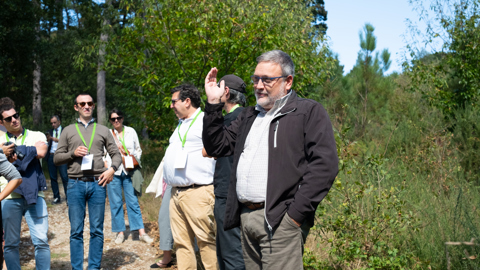 A group of people stands on a rocky path outdoors in a wooded area. A man in the foreground with gray hair and glasses raises his hand while others listen, some wearing lanyards and sunglasses. Bright, sunny weather.