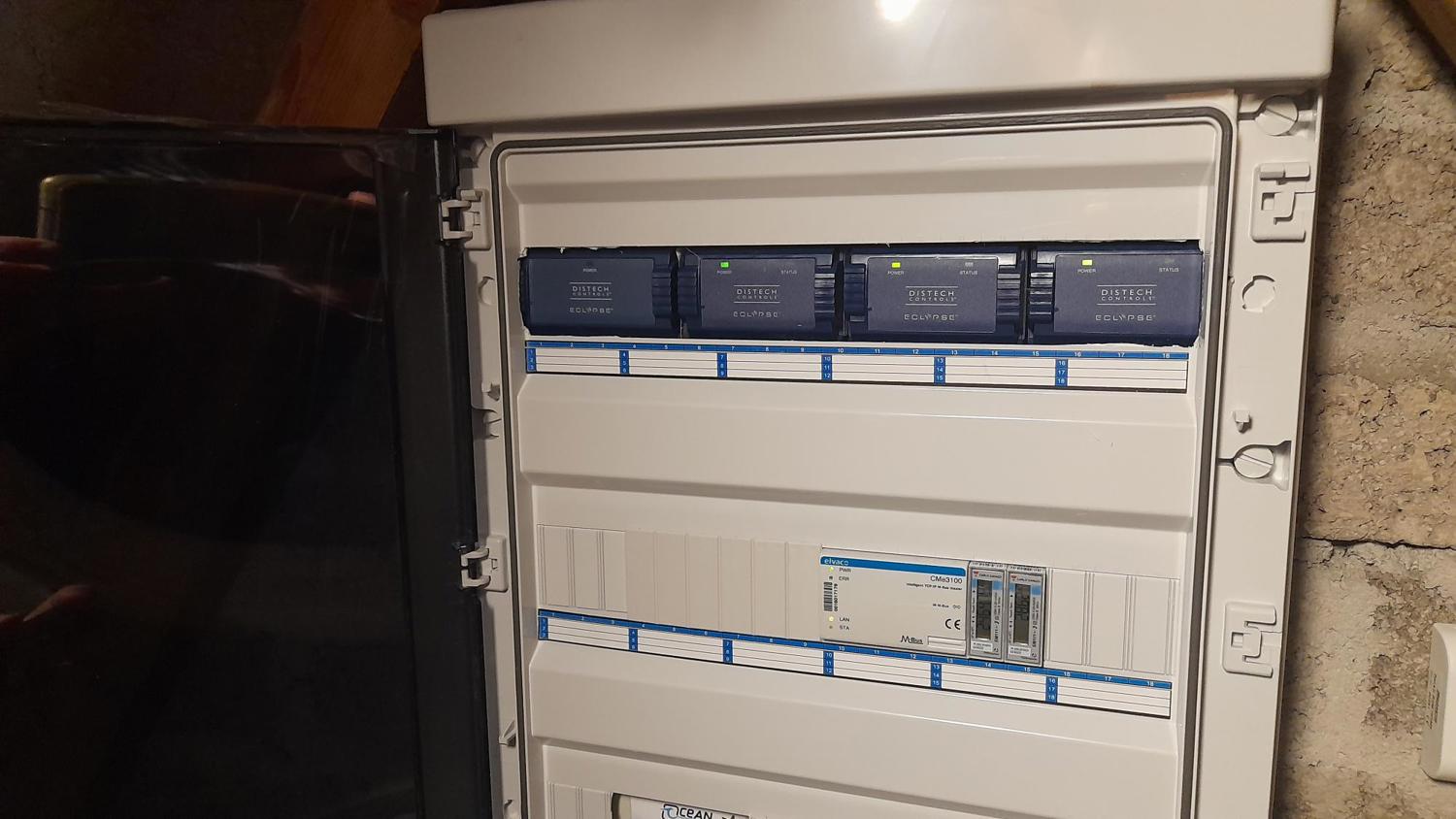 An open electrical panel mounted on a wall, showing several installed circuit breakers and devices, some with small green indicator lights illuminated. The panel cover is hinged open to the left.