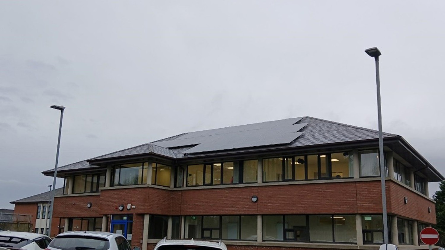 A two-story brick office building with large windows and solar panels on the roof. Several white cars are parked in front, and streetlights stand nearby under an overcast sky.