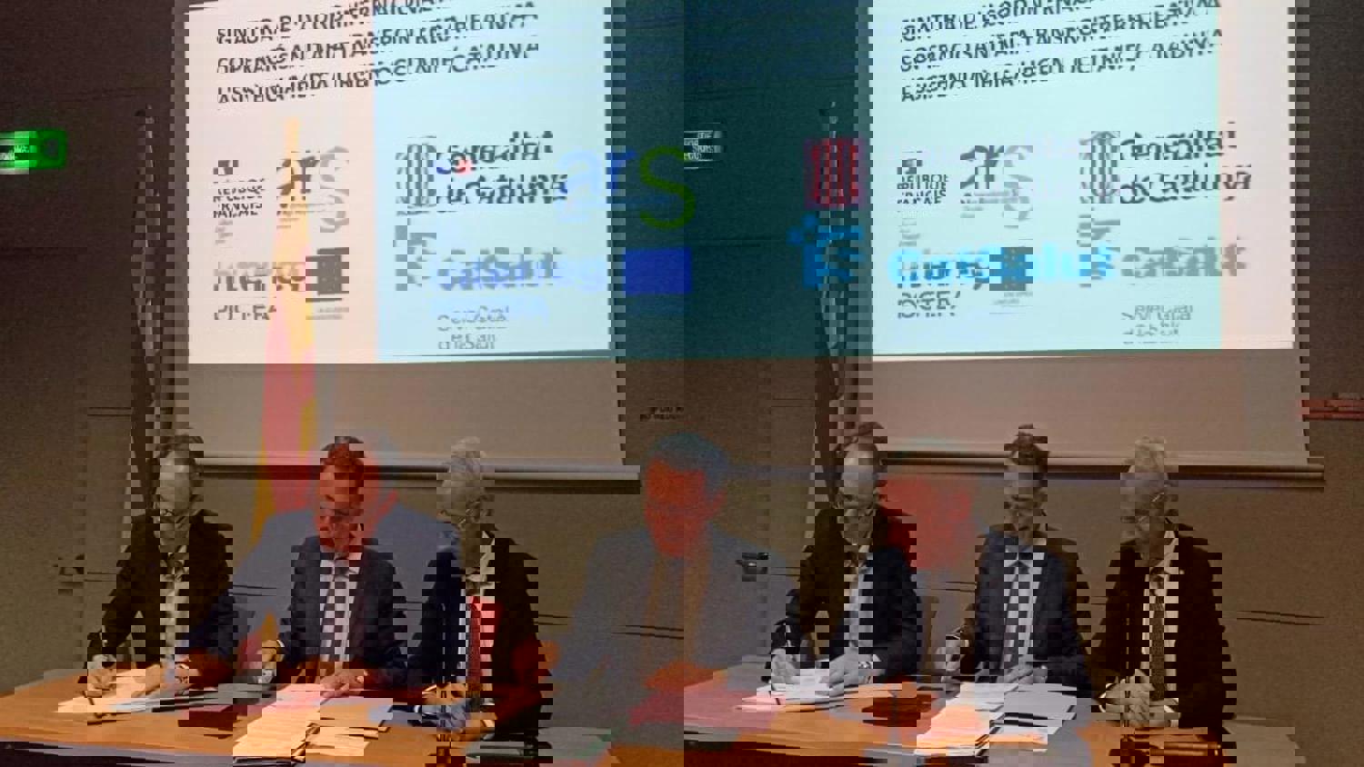 A distorted image shows several people seated at a panel in front of a projection screen displaying a presentation with various logos and text. The image is corrupted with horizontal glitches and digital artifacts.