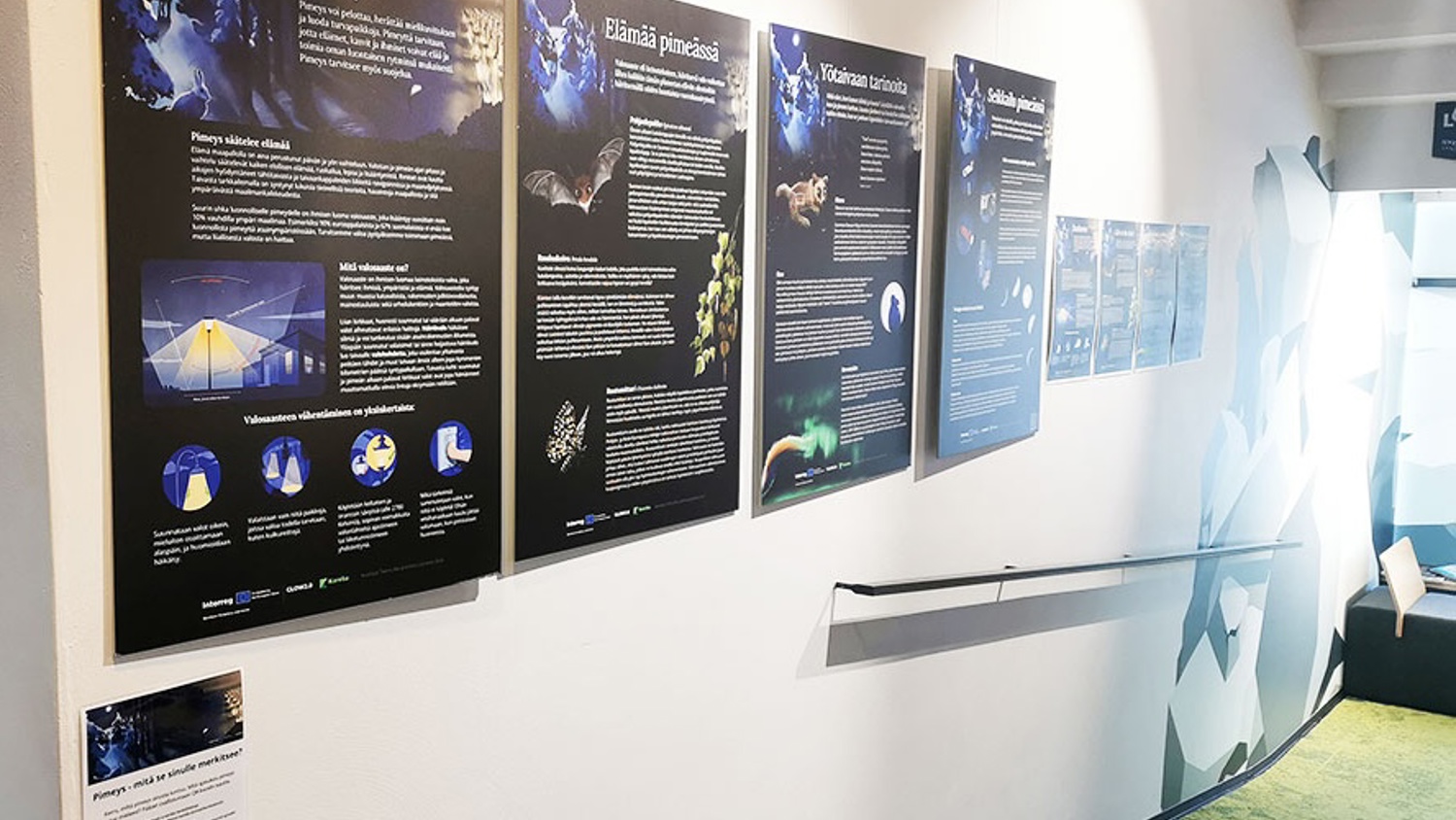 Exhibit wall featuring several informational posters in Finnish about darkness, animals, and nature, with images, diagrams, and a QR code; modern interior with a ramp and seating area visible.