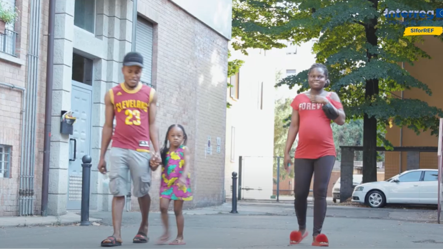 A man in a sports jersey and cap walks hand in hand with a child in a colorful dress. A woman in a red shirt and pants walks beside them on a paved path in an urban area with trees and buildings.