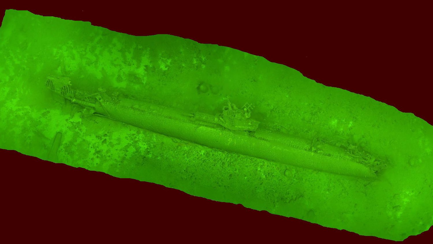 Green-tinted sonar image of a sunken submarine on the ocean floor. The submarine appears elongated and partially covered by sediment, surrounded by a dark background.