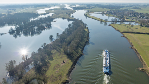 Aerial view of a river with a large white boat sailing, surrounded by green fields, patches of trees, and partially flooded land under a clear blue sky.