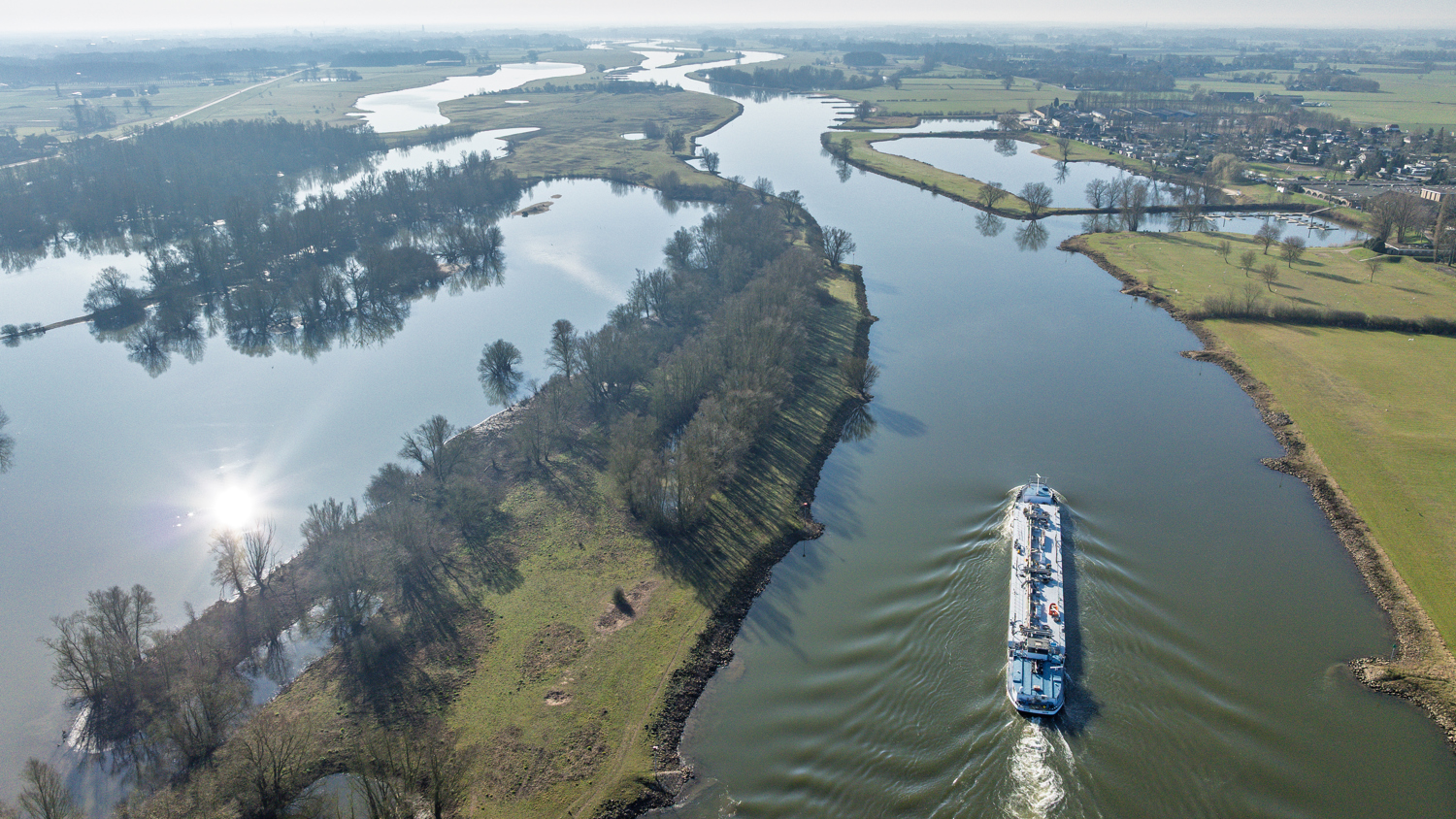 Aerial view of a river with a large white boat sailing, surrounded by green fields, patches of trees, and partially flooded land under a clear blue sky.