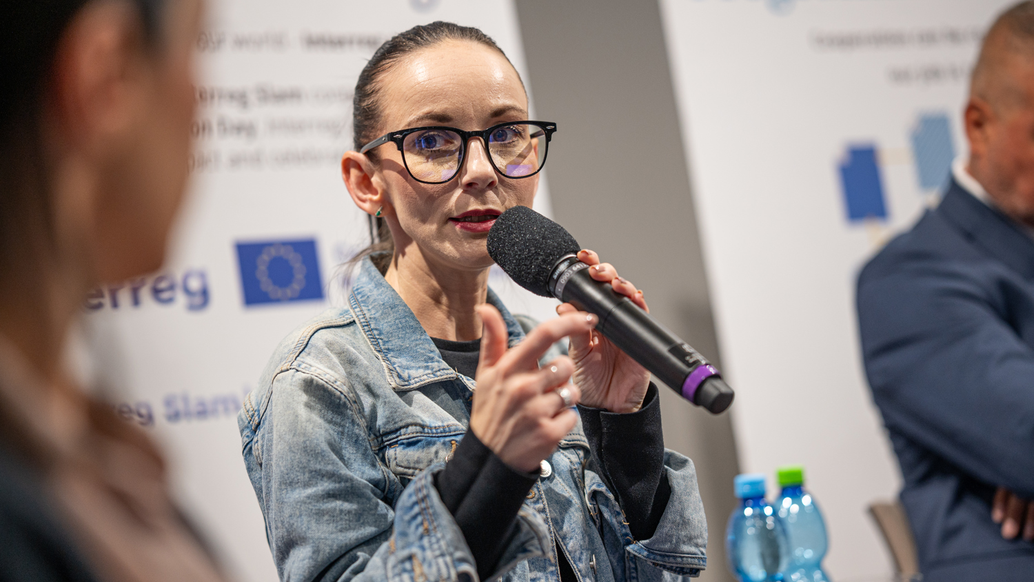 A woman wearing glasses and a denim jacket speaks into a microphone at a panel discussion. Two other people are partially visible, with banners and water bottles in the background.