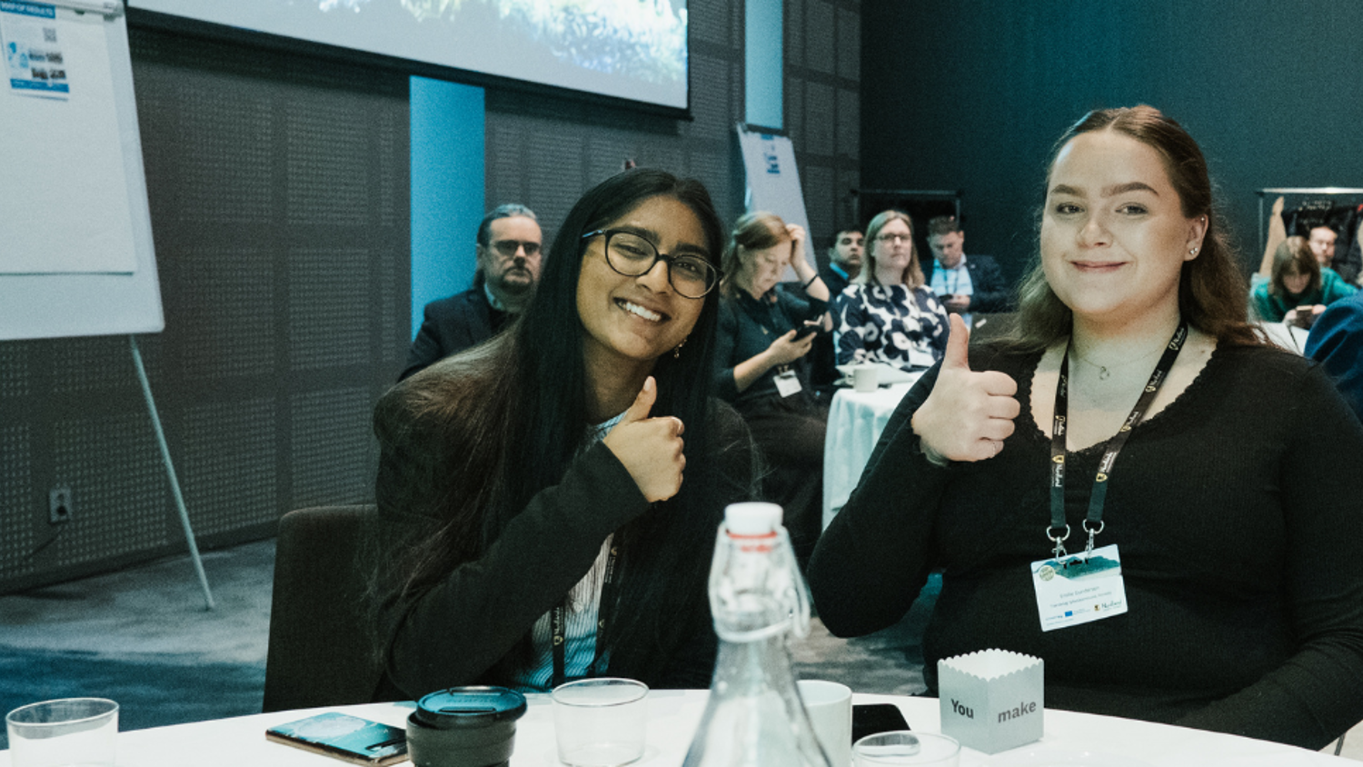 Two women sitting at a conference table smile and give a thumbs-up to the camera; other attendees can be seen seated in the background.