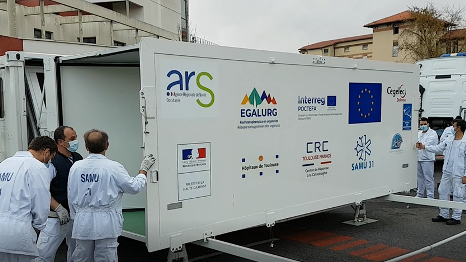 Several people in protective suits move a large white container featuring various logos, including ARS and European Union symbols, on a street surrounded by buildings and trucks.