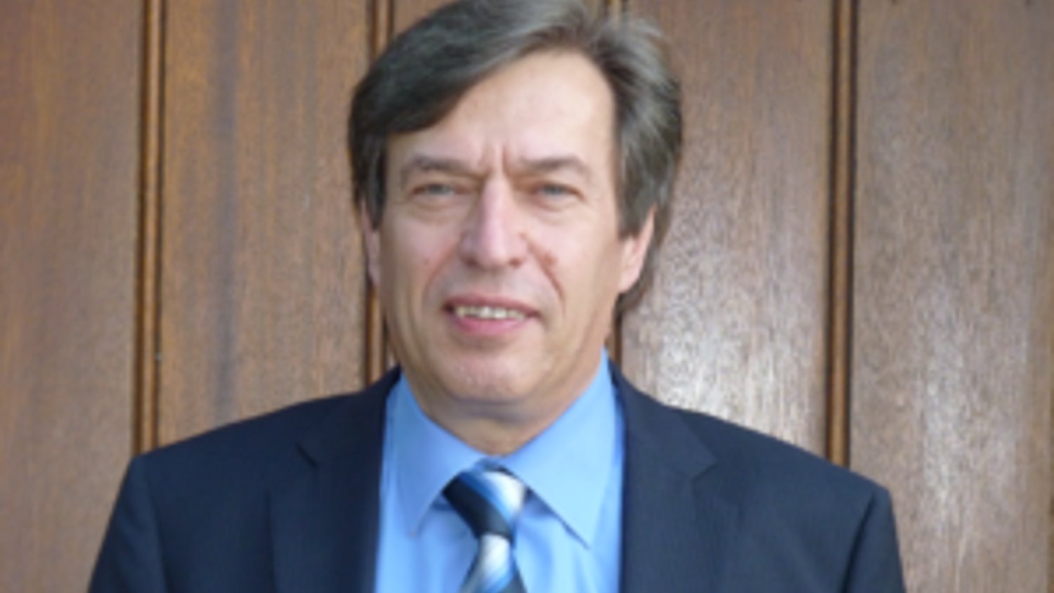 A person wearing a blue shirt and a striped tie with a dark suit stands in front of a wooden wall.