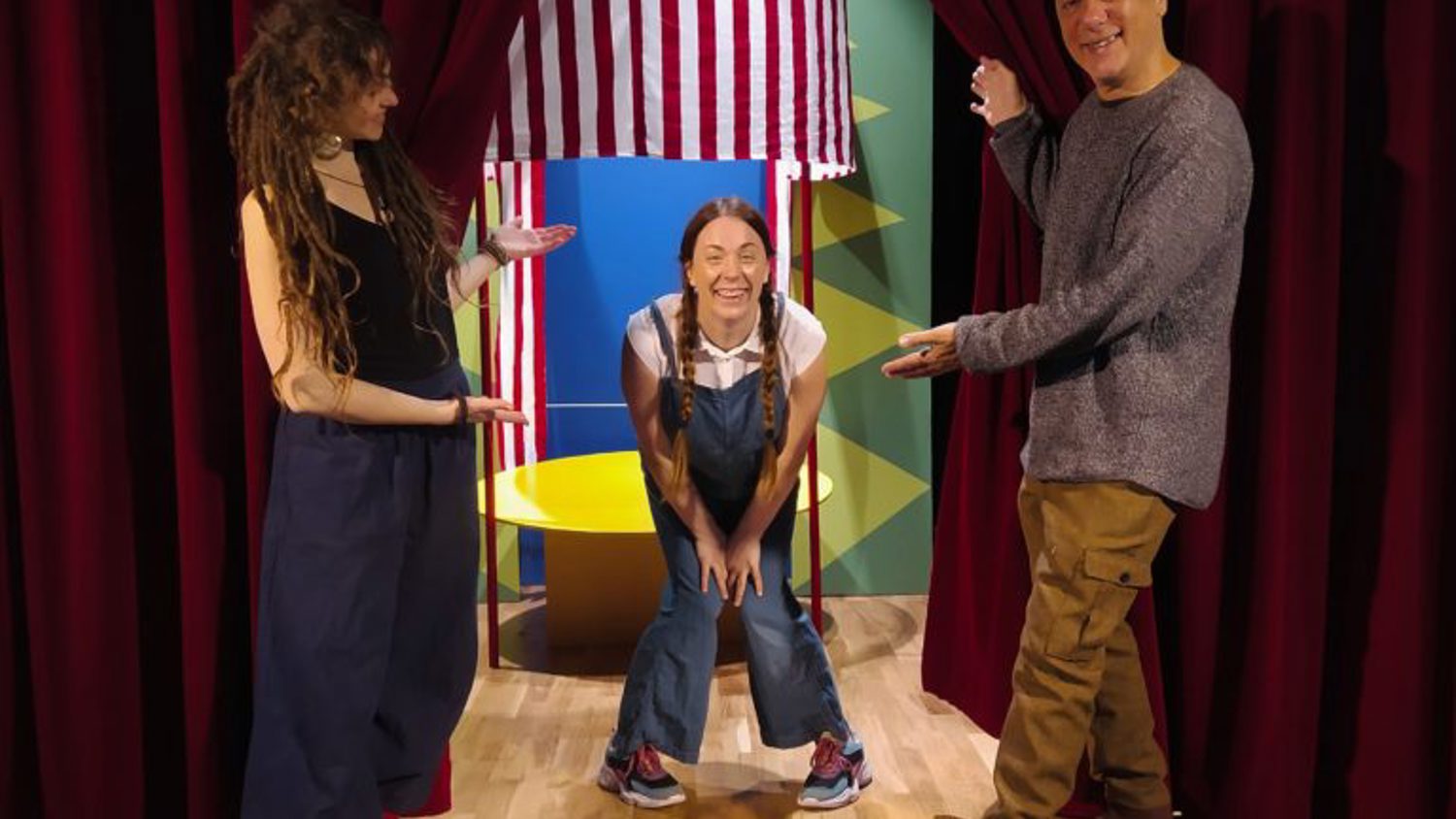 Three people pose on a stage with red curtains. A person in the center crouches smiling, wearing overalls and pigtails. The two others stand on either side, gesturing toward the center person. A striped background is partially visible behind them.