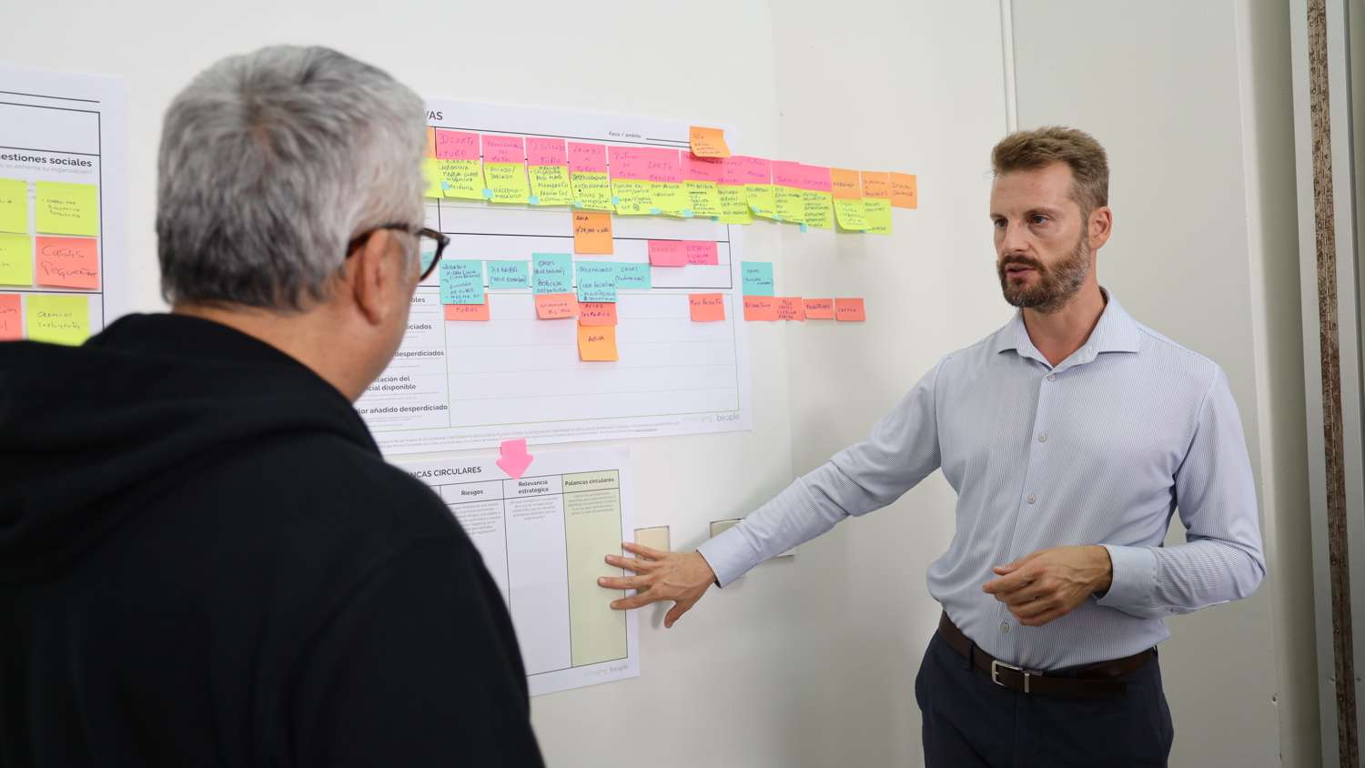 Two men discuss in front of a whiteboard covered with colorful sticky notes and charts. One man gestures toward the board while explaining something to the other, who listens attentively.