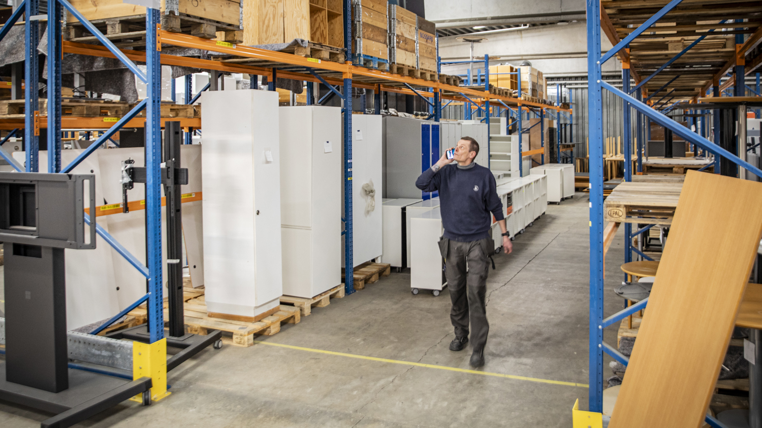 A man in a warehouse walks between racks of boxes and large white cabinets, talking on a mobile phone. The warehouse is organized with shelves, pallets, and industrial lighting overhead.
