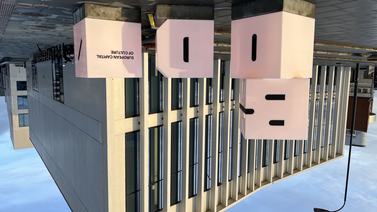 Large pink cubes with black letters and the words "European Capital of Culture" stand in front of a modern beige and glass building on a paved street under a blue sky.