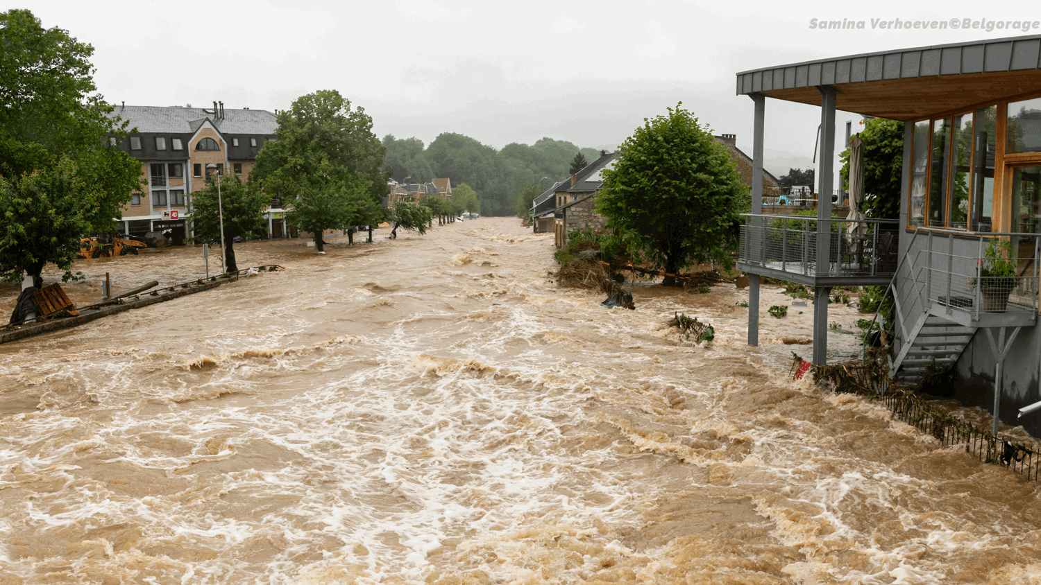 Floodwaters rush through a residential street, submerging roads and reaching building foundations. Trees and houses line the street under a cloudy sky, and debris is scattered in the turbulent water.