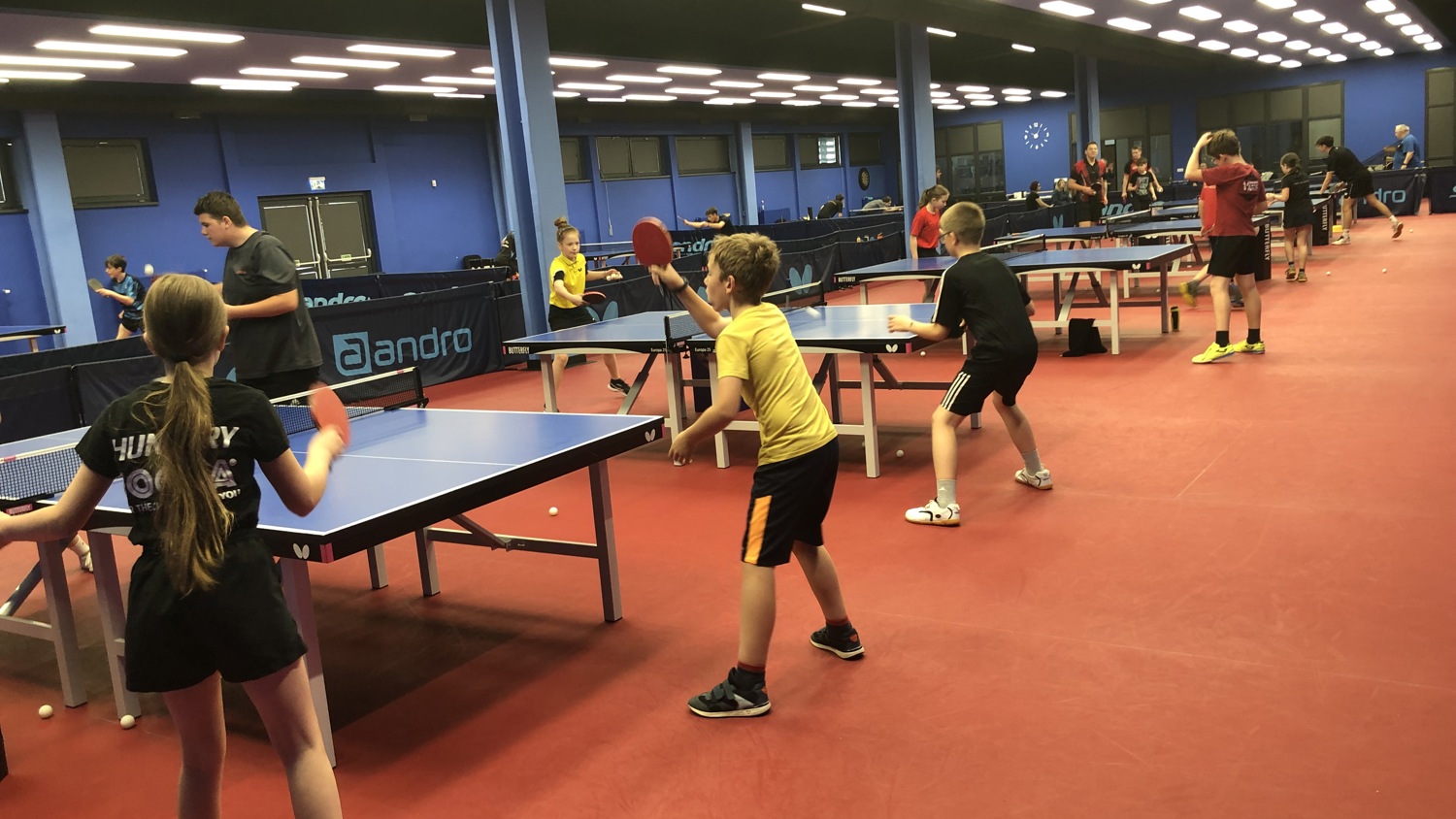 A group of people playing table tennis in an indoor facility with blue walls and bright overhead lights. Players are actively engaged, holding paddles, with several tables lined up. The floor is red, contributing to the sporty atmosphere.