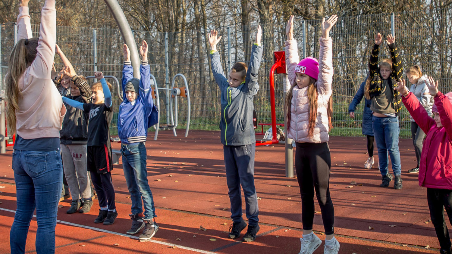 A group of children and an adult stand on a red outdoor sports court, with trees in the background. They are stretching, raising their arms upwards under a clear sky, engaging in a group exercise activity.