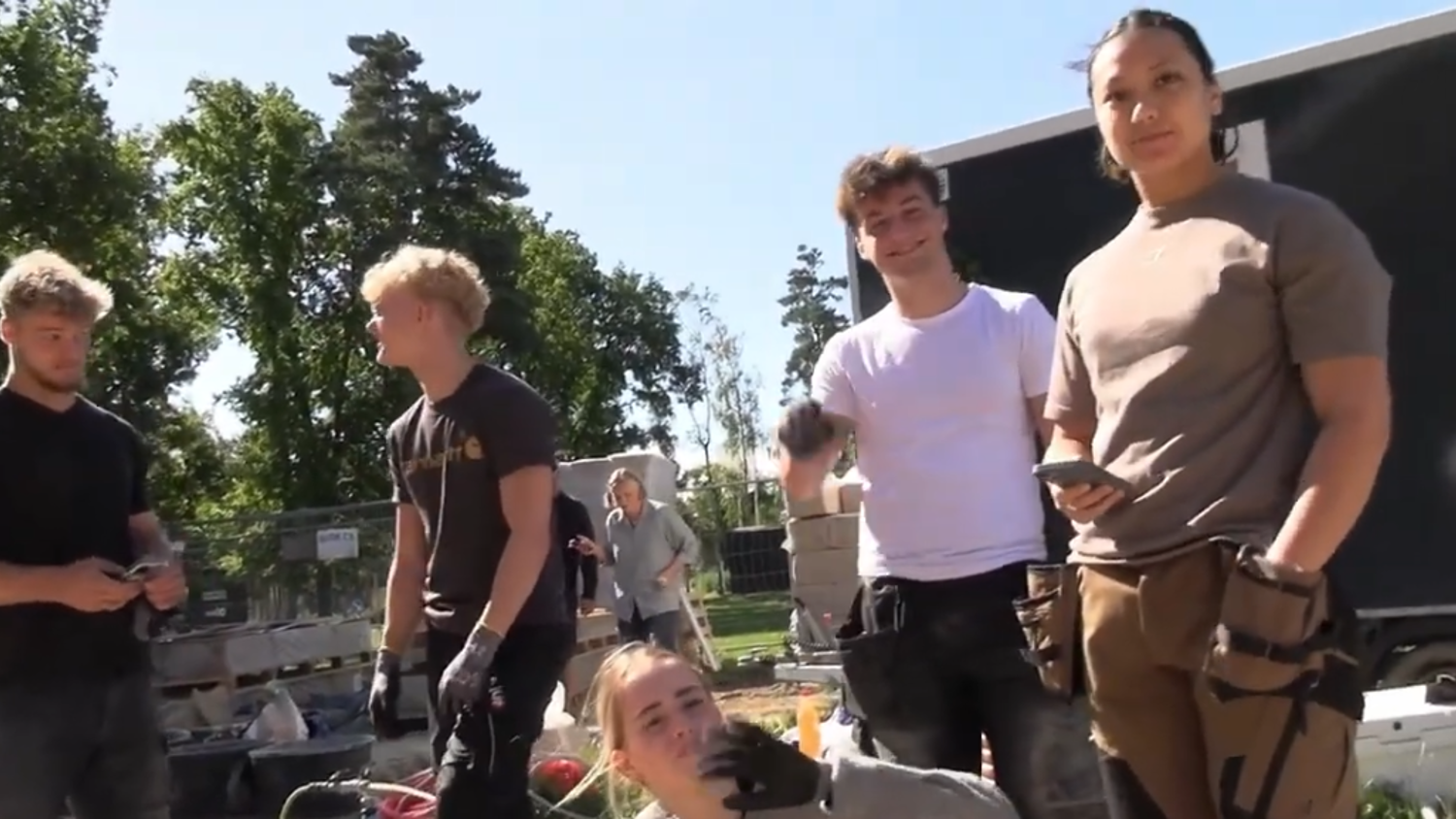 A group of five young adults wearing work clothes and gloves stand and kneel outdoors near construction materials and a building, with trees and clear skies in the background.