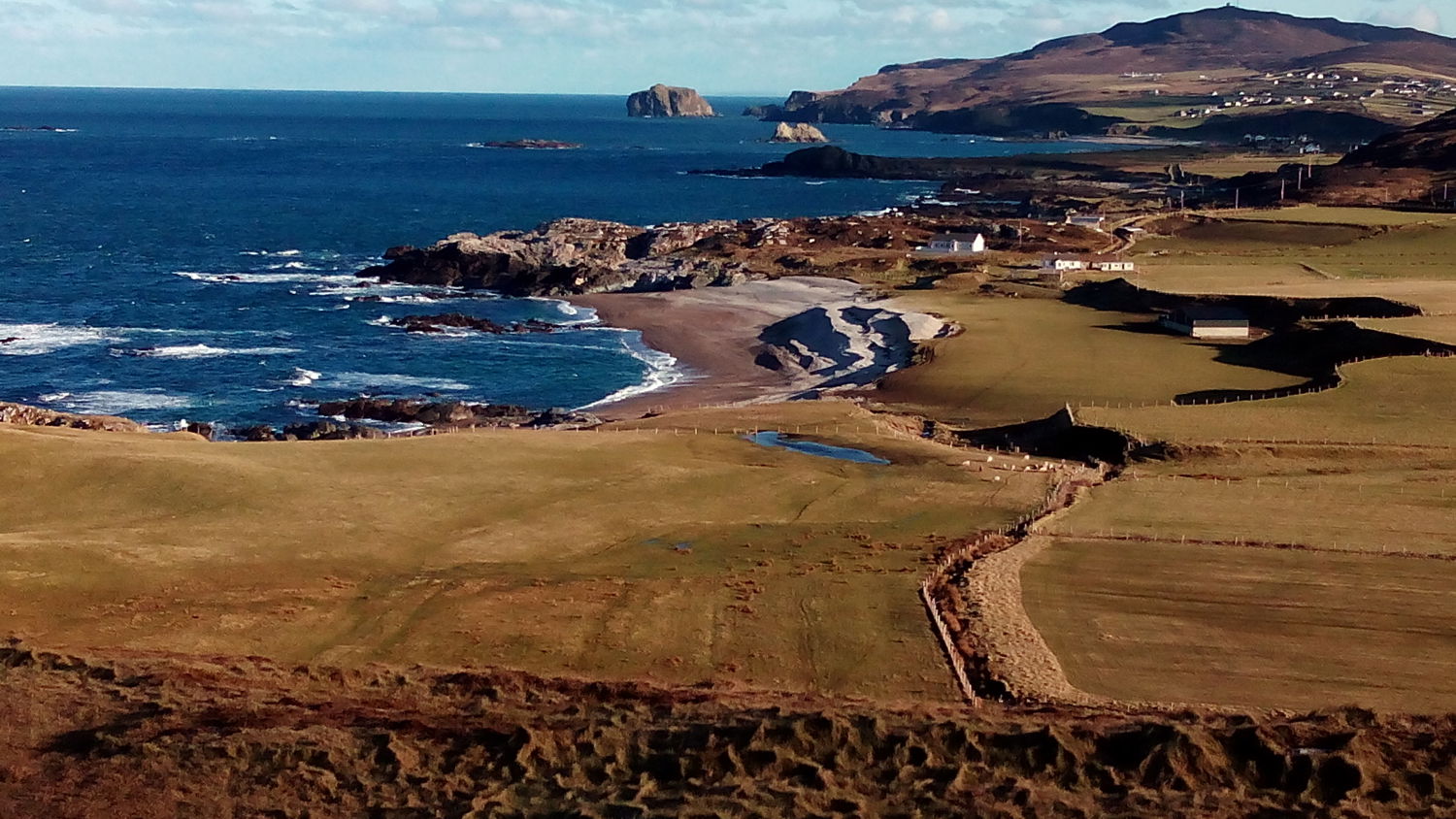 A coastal landscape with grassy fields, a curved sandy beach, rocky shoreline, and distant hills. Scattered houses and farm buildings are near the shore under a blue sky with light clouds.