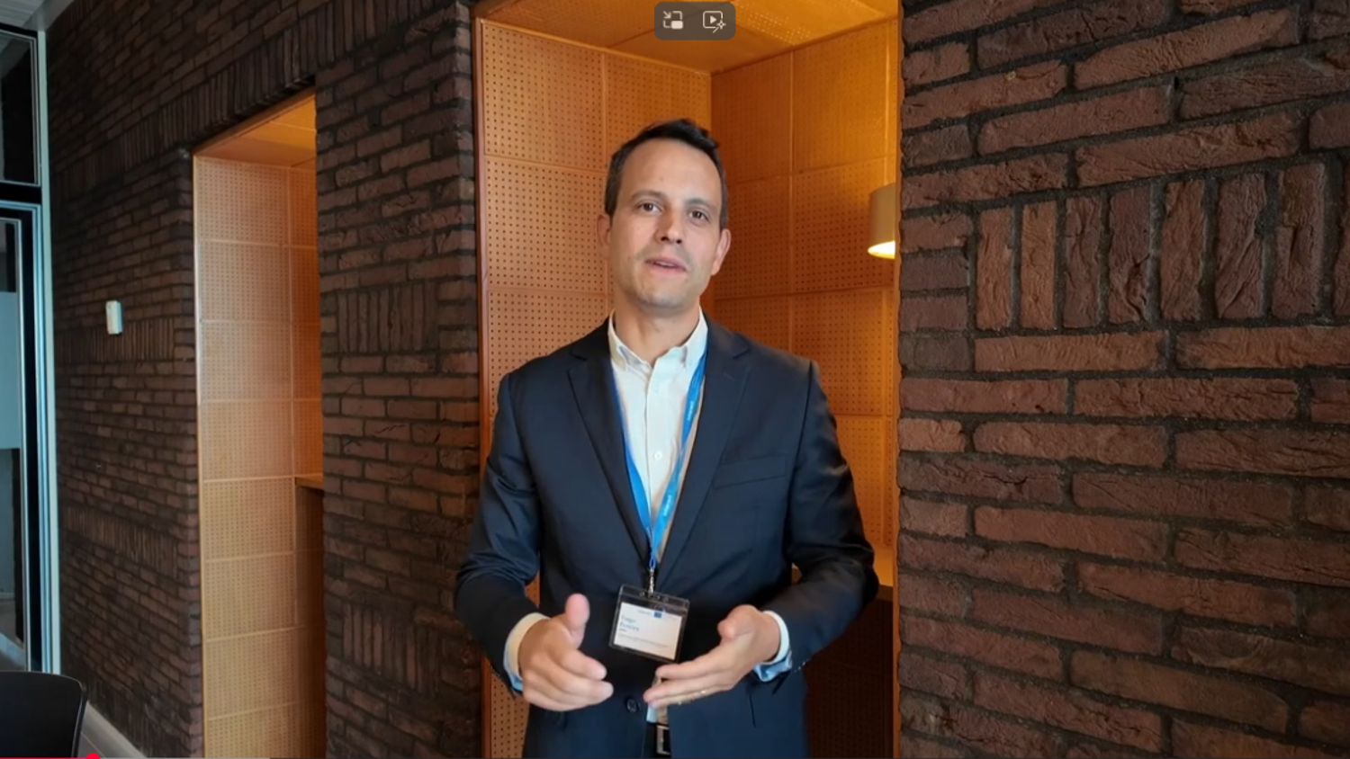 A man in a dark suit and white shirt stands indoors against a brick wall, wearing a lanyard and name badge, gesturing with his hands while looking at the camera.