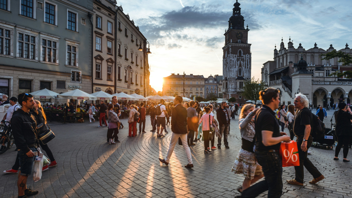 A bustling town square during sunset with people walking and socializing. Historic buildings and a clock tower are in the background. Shop awnings and outdoor seating are visible, with the golden light creating a warm atmosphere.