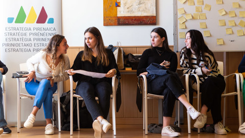 Four young women sit in chairs in a bright room, engaged in conversation. Behind them are a banner with multilingual text, a colorful painting, and a board covered with sticky notes.
