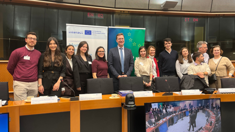 A group of people dressed in business attire stand together, smiling, in a conference room with banners, microphones, and a large screen showing a meeting in progress.