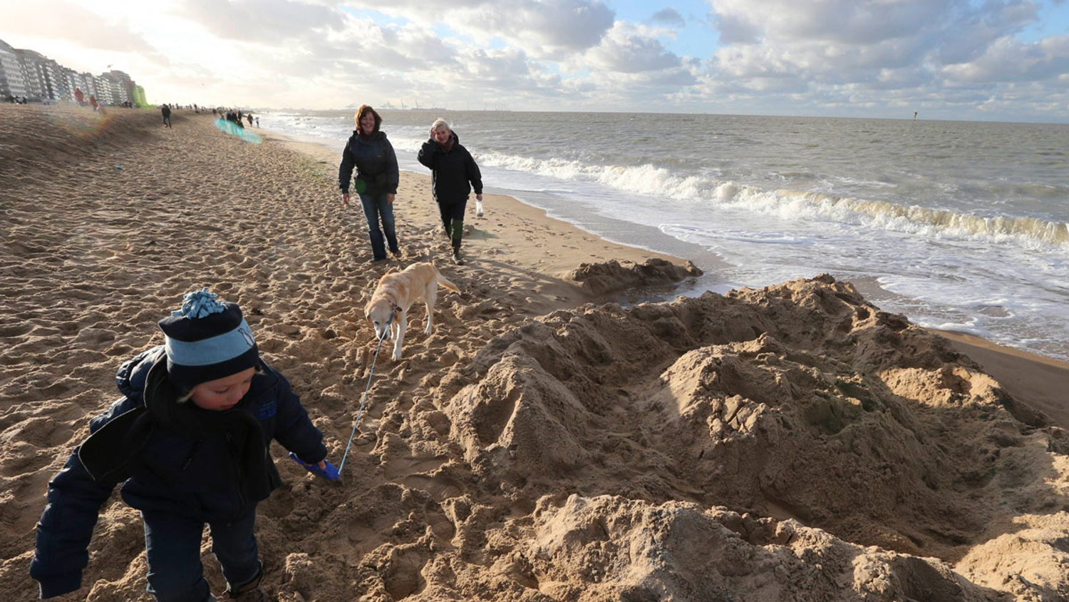 A child in a blue jacket plays on a sandy beach with a small shovel. Two adults walk a dog along the shoreline. The ocean waves crash gently, and buildings are visible in the distance under a cloudy sky.