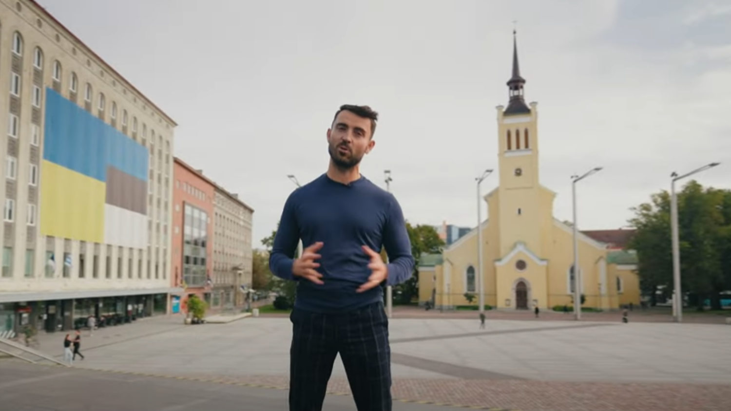 A man stands in a public square, gesturing with his hands. Behind him is a large building with a Ukrainian flag design on it, and a yellow church with a spire. The sky is overcast.