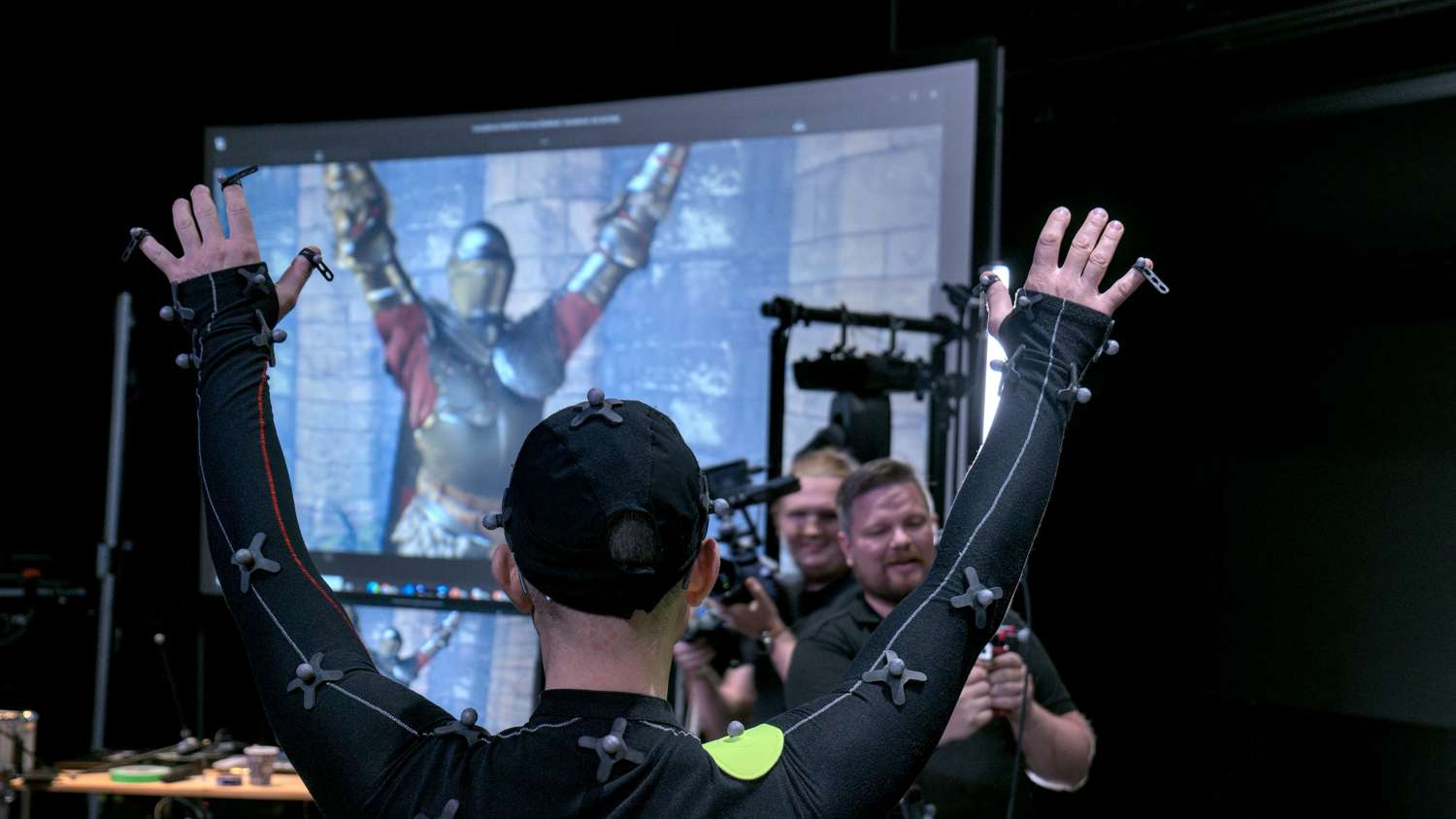 A person wearing a motion capture suit with sensors raises their arms in a studio, while crew members smile and work with equipment. A large screen behind displays an armored knight mirroring the same pose.