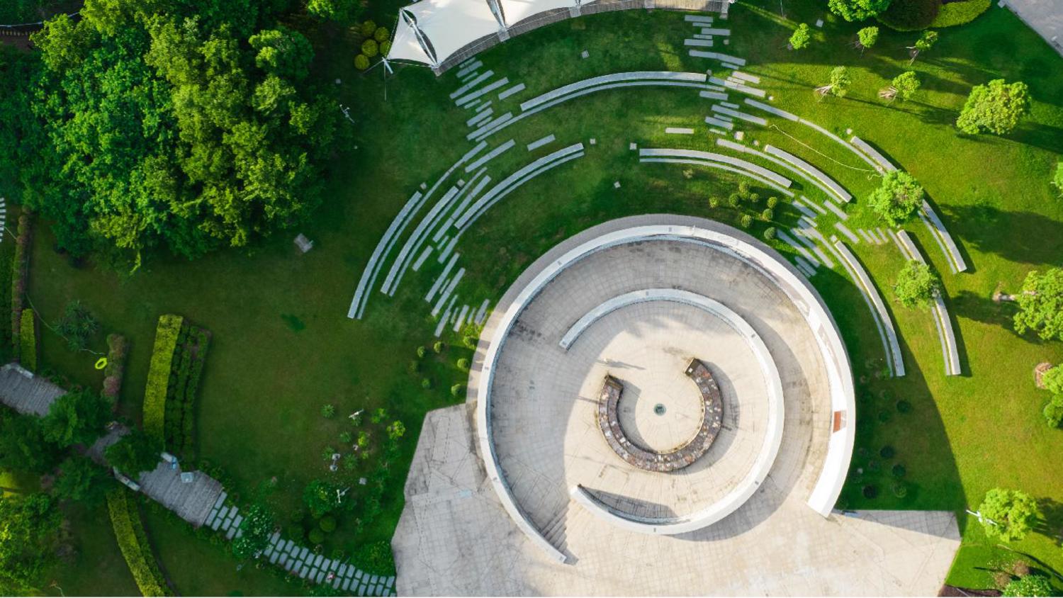 Aerial view of a circular stone plaza with concentric pathways and benches, surrounded by lush green lawns and trees on a sunny day. Curved paths and landscaping create an attractive geometric design.