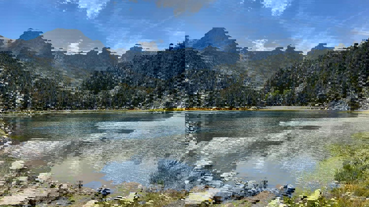 A scenic mountain lake with rocky hills and pine trees reflecting on the water under a bright blue sky, with some digital distortion visible across the image.