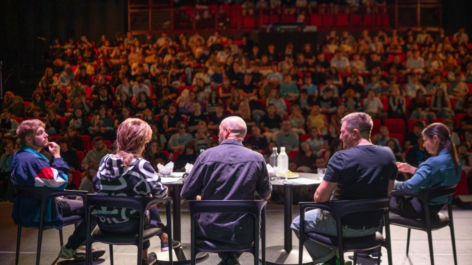 Five people sit on a stage, facing an audience in a crowded auditorium. The audience appears engaged, and the panelists are seated around a table with microphones and water bottles.