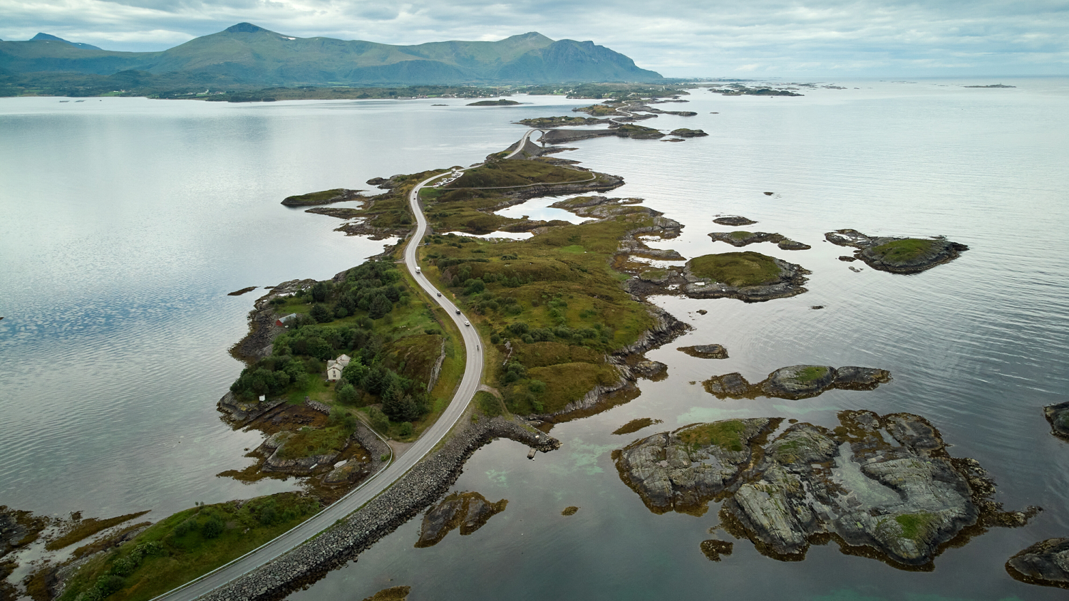A winding road stretches across small islands and rocky outcrops surrounded by calm water, with distant green mountains under a cloudy sky.