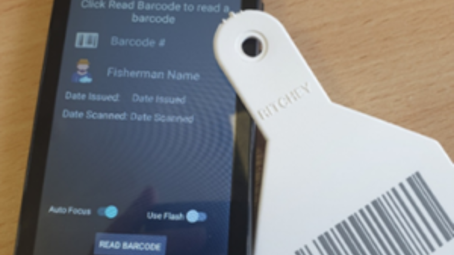 A smartphone on a wooden surface displays a barcode scanning app interface. Next to it is a white tag with a visible barcode and the number "0086.