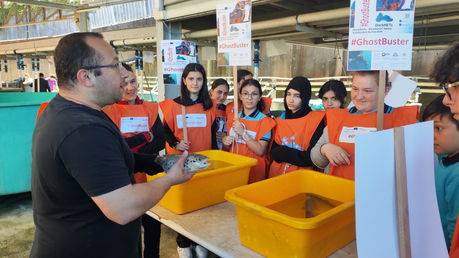 A man in a black shirt holds a fish over a yellow bin while a group of students in orange vests, holding “#GhostBuster” signs, watch intently in an indoor aquaculture facility.