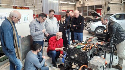 A group of people stand and kneel around a partially disassembled electric car engine in a workshop, listening to a person in a red shirt explain something. Various tools and car parts are visible.