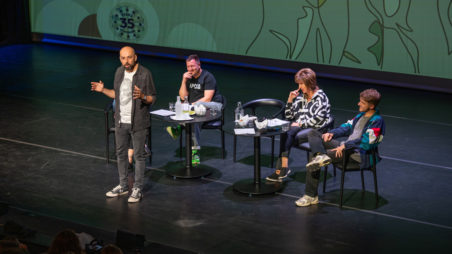 A man stands and speaks on stage with three people seated behind a table. A large screen with abstract designs is behind them. The audience is visible in the foreground.
