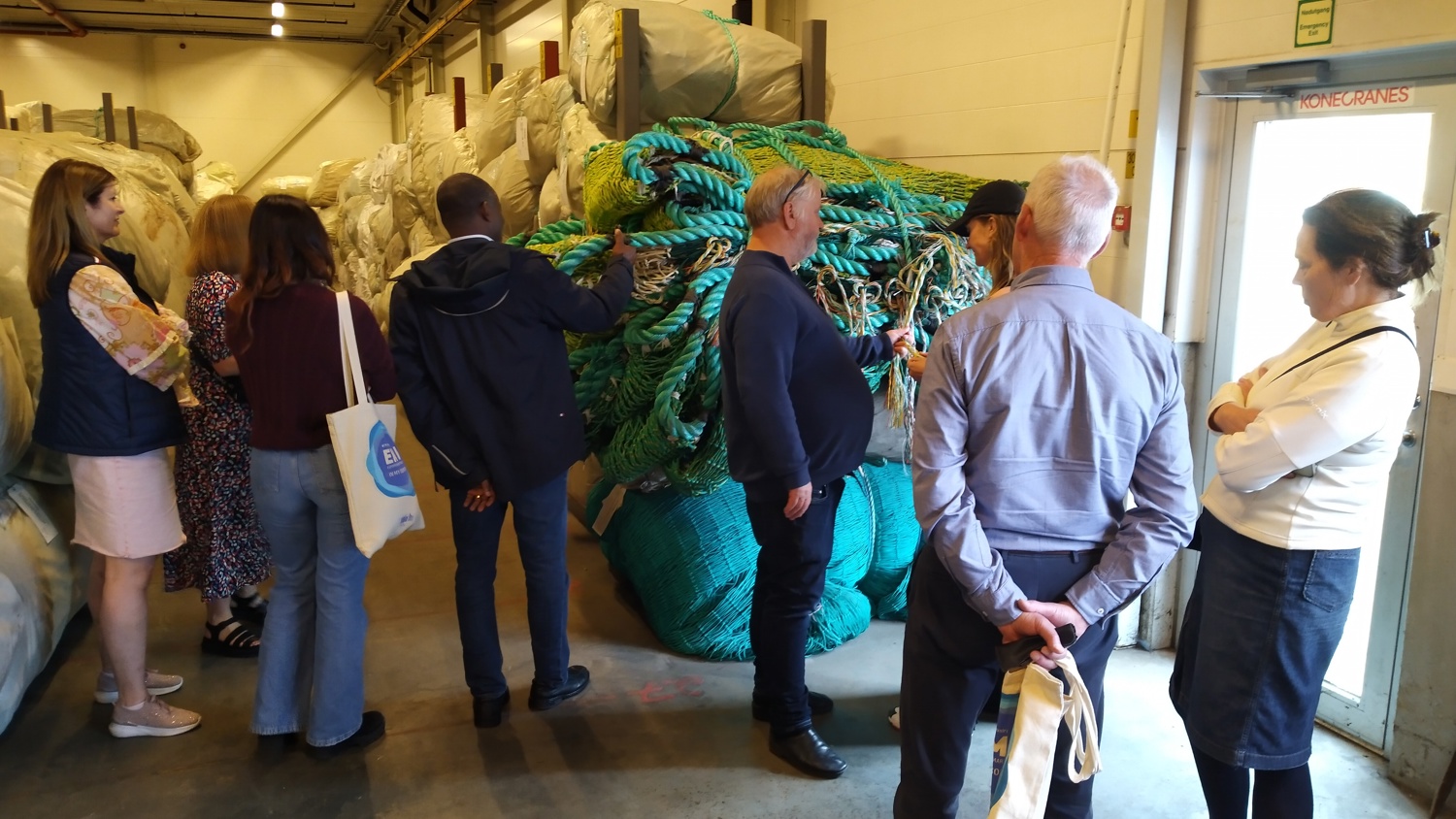 A group of people stands around a large bundle of colorful ropes in a warehouse. Some are examining the ropes, while others are observing. The space is filled with various large bags and containers. A labeled fire exit door is visible in the background.