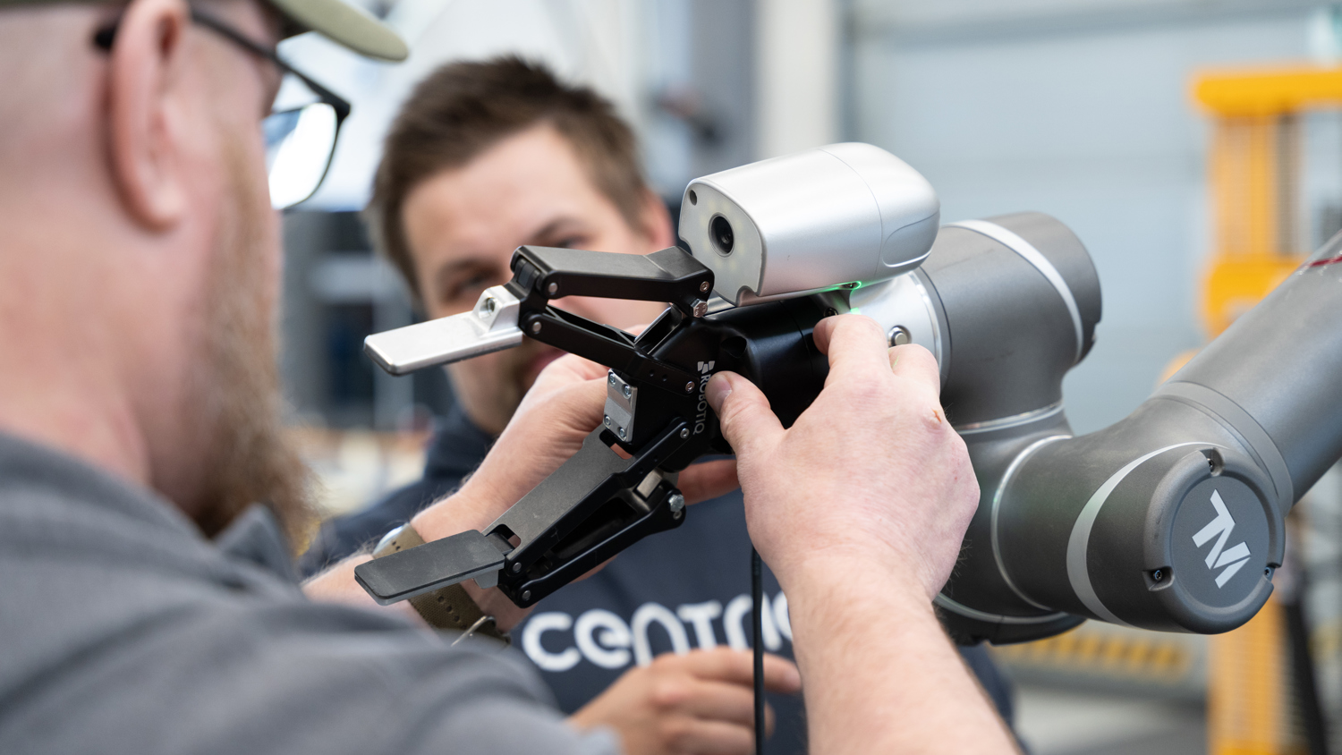 Two people work together, adjusting a robotic arm with a mechanical gripper in a modern industrial setting. One person holds the gripper while the other observes. The atmosphere is collaborative and technical.