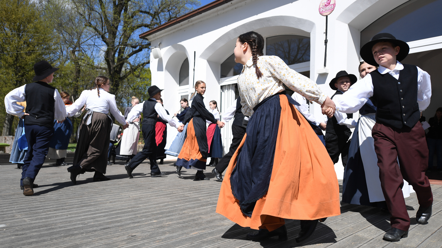 A group of people in traditional folk costumes dance in a circle on a wooden stage outdoors. The women wear long skirts and blouses, while the men wear vests and hats. It's a sunny day with trees in the background.
