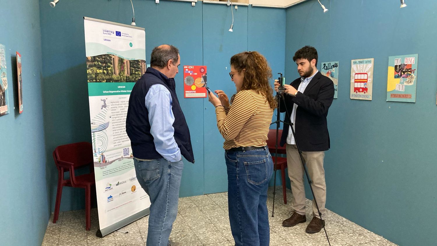 A woman with curly hair interviews a man in a blue vest against a teal wall, while a man in a suit films the interaction. The room is decorated with colorful posters and a tall banner.