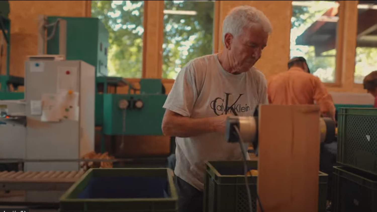 An older man in a Calvin Klein shirt works in a workshop or factory setting. He is using a piece of equipment with various bins and machinery visible in the background. Natural light streams in through large windows.