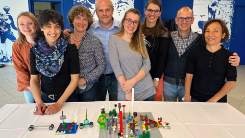 Eight smiling adults stand behind a table displaying colorful LEGO creations on a white tablecloth, in a bright room with blue and white walls.