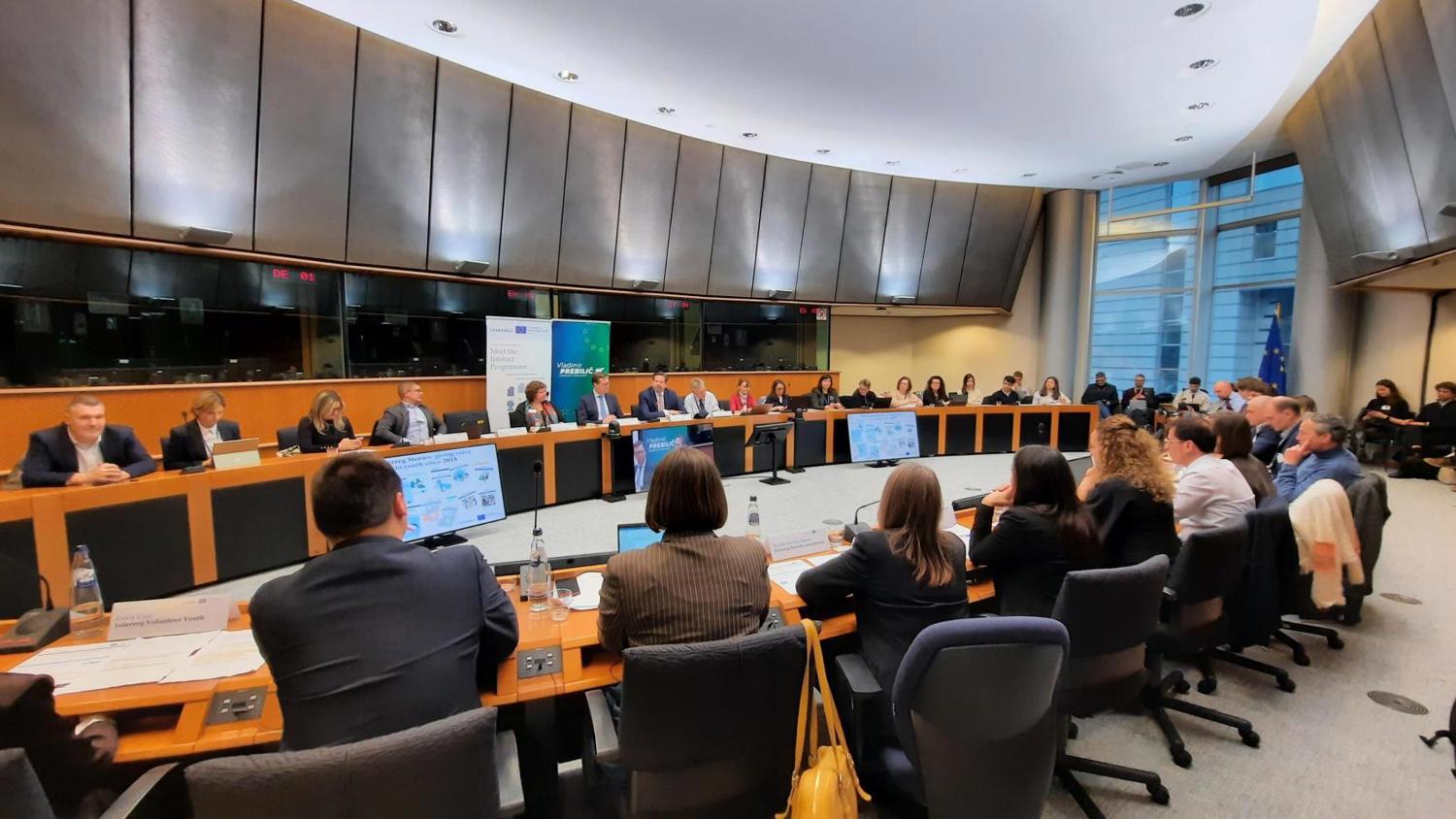 A large group of people sit around a curved conference table in a modern meeting room, engaged in discussion. Multiple screens and papers are visible, and large windows allow natural light to enter the space.